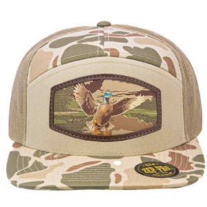Puede incluir: Gorra de camionero con estampado de camuflaje y panel frontal beige con un parche de cuero. El parche representa un pato en vuelo sobre un paisaje. La gorra tiene una visera curva y una parte trasera de malla.