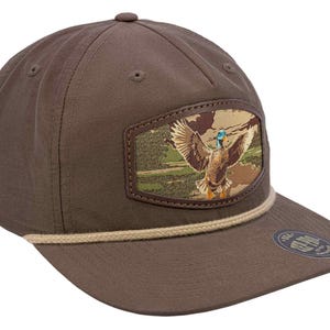 Puede incluir: Gorra de béisbol marrón con visera plana y un parche con un pato en vuelo. El parche tiene un fondo de camuflaje y un borde marrón. Un detalle de cuerda beige adorna la visera.