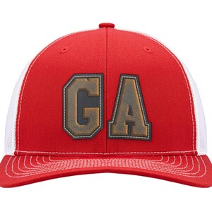 May include: A red baseball cap with white mesh back panels and a curved brim featuring white stitching. The front displays the letters GA in a large, block-style font with a dark, weathered, metallic-looking finish and a dark border outline.