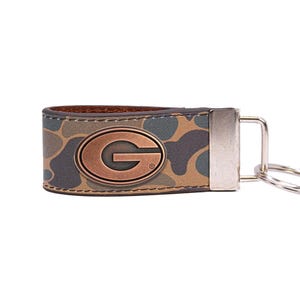May include: A leather keychain with a camouflage pattern and a copper-colored "G" emblem. The keychain has a silver-colored metal clasp and a key ring. The overall color scheme is brown, green, and gray.