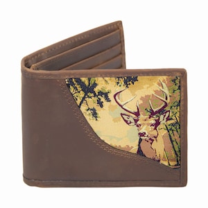Puede incluir: Cartera marrón de dos pliegues con un gráfico de ciervo en la parte delantera. La cartera presenta una ilustración detallada de un ciervo en un entorno forestal, con tonos de marrón, verde y beige. El interior tiene ranuras para tarjetas.