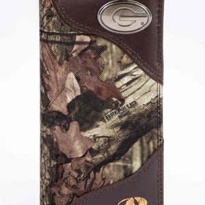 Puede incluir: Una cartera de tres pliegues con un patrón de camuflaje, detalles en cuero marrón y un emblema plateado con la letra "G". La cartera presenta los logotipos "BREAK UP INFINITY" y "MOSSY OAK". La cartera está hecha de tela y cuero.