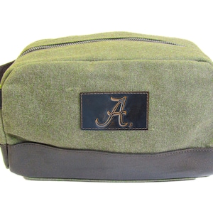 以下が含まれることがあります： 緑色のキャンバス地の化粧ポーチで、茶色の革の縁取りと、茶色の革のパッチに筆記体で「A」の文字が書かれています。