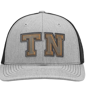 May include: A heather grey baseball cap with a black mesh back and a structured crown. The front features the letters TN in a bold, block-style font with a dark brown, leather-like texture and a dark outline, centred on the grey fabric panel.