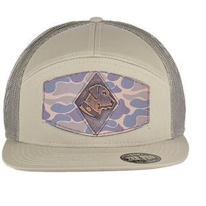 Könnte beinhalten: Eine beige und graue Trucker-Kappe mit einem Tarnmuster und einem Leder-Patch mit einem Hundekopf. Die Kappe hat ein Netzrücken und eine flache Krempe.