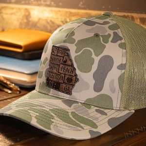May include: A camouflage trucker hat with a mesh back and a curved brim. The front panel features a leather patch in the shape of the state of Georgia, with text and a peach graphic. The hat is in shades of green, gray, and tan.