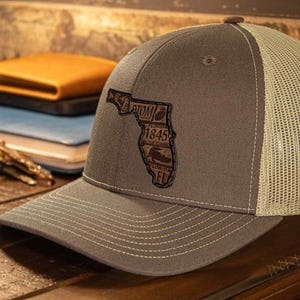 May include: A gray and tan trucker hat with a stitched brim and a mesh back. The hat features a brown patch in the shape of Florida with the text "ATOM 1845 FL". The hat is sitting on a wooden surface.