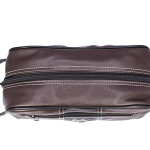 Puede incluir: Un neceser de cuero marr&oacute;n con cremallera negra y costuras blancas. La bolsa tiene un asa en un lado y un peque&ntilde;o bolsillo con cremallera en la parte delantera. La bolsa es de forma rectangular.