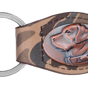 May include: A brown leather keychain with a camouflage pattern. It features a metal oval charm with a copper-colored dog head.