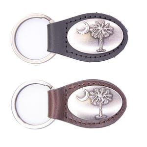 May include: Two keychains featuring the South Carolina state flag emblem. Each keychain has a leather strap in either black or brown, with a silver-colored oval plate displaying a crescent moon and a palmetto tree. The key rings are silver.