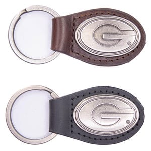 Op de afbeelding: Twee leren sleutelhangers, één bruin en één zwart, elk met een zilverkleurig ovaal embleem met de letter "G". De sleutelhangers hebben een metalen ring om sleutels aan te bevestigen.