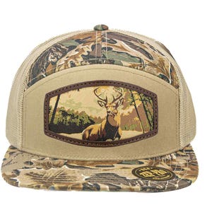 Pode incluir: Boné de caminhoneiro bege e camuflado com um patch retangular com um veado em uma cena de floresta. O boné tem uma parte traseira de malha e uma aba curva com um padrão de camuflagem.