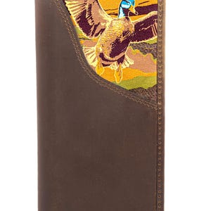 Puede incluir: Cartera de cuero marrón con un diseño de pato bordado colorido. El pato se representa en vuelo con las alas extendidas, sobre un fondo de tonos tierra. La cartera tiene un diseño plegable.
