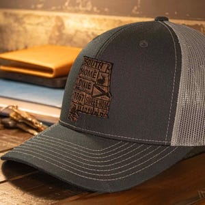 May include: A charcoal gray trucker hat with a gray mesh back. The front of the hat features a brown leather patch with the words "South Home Dixie 1867 Sweet Home Alabama" in a map outline.