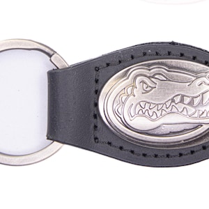 May include: Black leather keychain with a silver-colored oval emblem featuring a detailed alligator design. The keychain includes a silver-colored split ring for attaching keys. The emblem is stitched onto the leather.