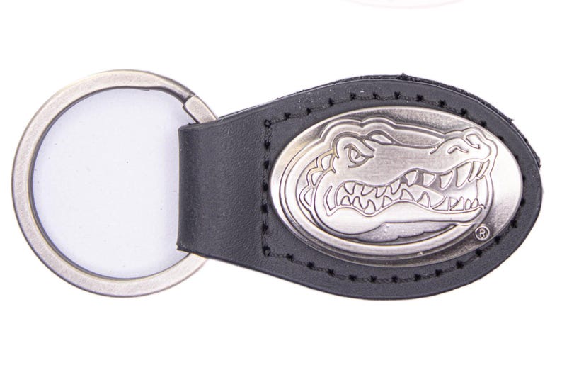 May include: Black leather keychain with a silver-colored oval emblem featuring a detailed alligator design. The keychain includes a silver-colored split ring for attaching keys. The emblem is stitched onto the leather.