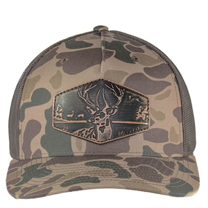 Puede incluir: Una gorra de camionero de camuflaje con un parche de cuero marrón que presenta una silueta de ciervo. El parche tiene un aspecto desgastado y está cosido en la parte delantera de la gorra.