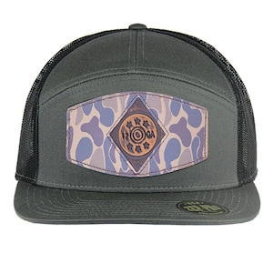 May include: A dark green trucker hat with a black mesh back. The hat features a camouflage patch with a brown leather circle that says "12 GA" in raised letters.