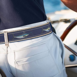 May include: A navy blue belt with a repeating pattern of sailfish in shades of blue, yellow, and green. The belt is worn with light-colored pants and a navy blue shirt. The belt is a nautical accessory.