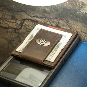 May include: A brown leather wallet with a magnetic money clip, holding two US dollar bills. The wallet features a silver emblem with the letters "OU". The wallet is resting on a black phone case and a blue leather surface.