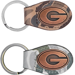 May include: Two keychains with a camouflage pattern leather tag and a copper-colored oval emblem with the letter "G". The keychains have a silver key ring attached to the leather tag.