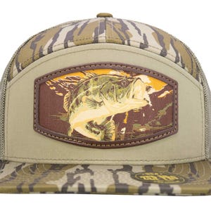 May include: A baseball cap with a camouflage pattern and a mesh back. It features a rectangular patch with a fish illustration, framed by a brown border. The fish is depicted in green, yellow, and brown hues.