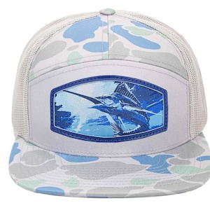 May include: A light grey trucker hat with a blue and white camouflage pattern on the brim and top. The front panel features a blue and white image of a sailfish. The hat has a mesh back and a curved brim.