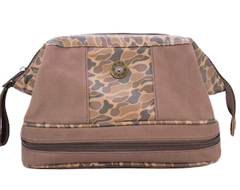 Neceser Shot Shell para hombre. Lona encerada marrón con acabado de camuflaje clásico. Logotipo de cobre antiguo.