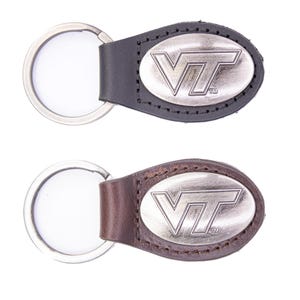 Op de afbeelding: Twee leren sleutelhangers met metalen ringen. De ene sleutelhanger is zwart, de andere bruin. Elke sleutelhanger heeft een ovaal metalen embleem met de letters "VT" in zilver. De sleutelhangers zijn waarschijnlijk gerelateerd aan Virginia Tech.