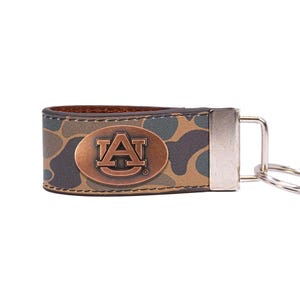 May include: A leather keychain with a camouflage pattern. The keychain features a bronze-colored oval emblem with the letters "A" and "U" inside. A silver-colored metal clasp and key ring are attached to the keychain.