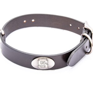 May include: A black leather dog collar with a silver buckle and a silver oval emblem featuring the letters "SC". The collar has a D-ring for leash attachment and is designed for a pet.