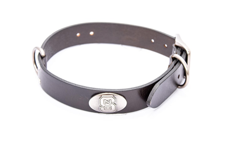 May include: A black leather dog collar with a silver buckle and a silver oval emblem featuring the letters "SC". The collar has a D-ring for leash attachment and is designed for a pet.