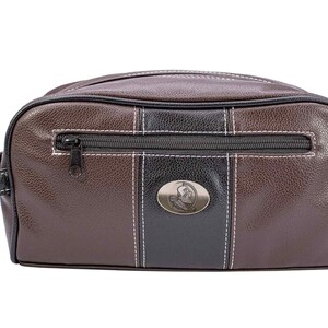 May include: A brown and black leather wash bag with a zip closure and a handle. The bag has a black stripe down the centre with a silver emblem and white stitching. The bag is rectangular and has a textured appearance.