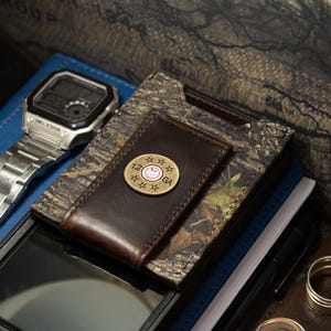 May include: A brown leather money clip with a decorative brass emblem featuring the text "12 GA" and a smiling face. The clip rests on a camouflage patterned wallet, with a silver wristwatch and a blue notebook in the background.