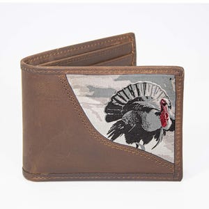 Puede incluir: Cartera de cuero marrón con un diseño de pavo en la parte delantera. La cartera tiene un diseño plegable con ranuras para tarjetas visibles en el interior. La ilustración del pavo utiliza tonos de gris, negro y rojo. La cartera es de cuero.