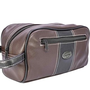 Puede incluir: Neceser de cuero marr&oacute;n con detalles en negro y blanco. Cuenta con una cremallera negra, asa y un emblema decorativo. La bolsa tiene forma rectangular y est&aacute; dise&ntilde;ada para viajar.