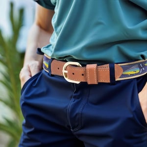 May include: A brown leather belt with a gold buckle. The belt features a navy blue fabric section with a fish pattern in yellow and blue. The belt is worn with navy blue pants and a teal shirt.