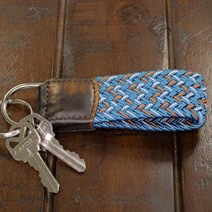 Op de afbeelding: Een sleutelhanger met een blauw, wit en oranje geweven ontwerp, bevestigd aan een leren band en metalen sleutelringen. Twee sleutels zijn bevestigd. De sleutelhanger ligt op een donkerbruin houten oppervlak.