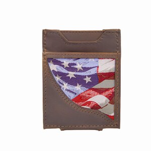 Puede incluir: Tarjetero de cuero marrón con un inserto de tela con el diseño de la bandera estadounidense. La cartera tiene forma rectangular con un borde cosido y un recorte en la parte superior.