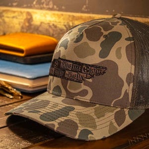 May include: Camouflage baseball cap with a dark gray mesh back. The front of the cap features a patch with the text "Tennessee State" and "Smoky Home In". The cap is sitting on a wooden surface.