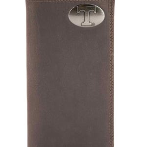 May include: A brown leather wallet with a silver oval emblem featuring a capital "T". The wallet is rectangular and has a stitched border. The wallet is closed and has a vertical design.