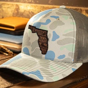 May include: A camouflage-patterned baseball cap with a mesh back and a curved brim. The front of the cap features a leather patch in the shape of Florida, with text and a logo. The cap is on a wooden surface.