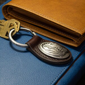 May include: A brown leather wallet and a key ring with a silver alligator emblem rest on a blue surface. The key ring has a brown leather strap and a silver metal ring. A set of keys is next to the key ring.