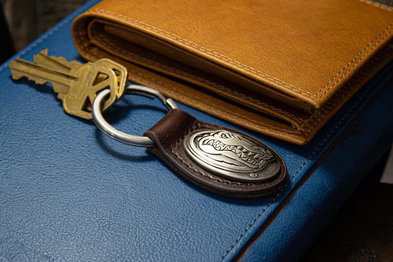 May include: A brown leather wallet and a key ring with a silver alligator emblem rest on a blue surface. The key ring has a brown leather strap and a silver metal ring. A set of keys is next to the key ring.