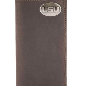 Puede incluir: Una cartera de cuero marrón con un emblema ovalado plateado con las letras "LSU". La cartera rectangular tiene un borde cosido. La cartera está cerrada y tiene un diseño simple y clásico.