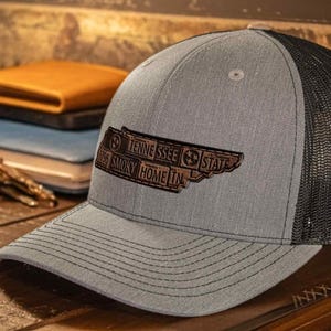 May include: Gray trucker hat with a black mesh back. The hat features a brown leather patch with the text "Tennessee State" and "Smoky Home In". The hat is sitting on a wooden surface.