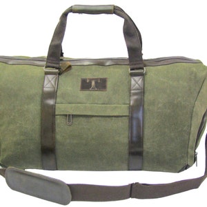 Puede incluir: Una bolsa de lona verde con detalles en cuero marrón y un bolsillo con cremallera en la parte delantera. La bolsa tiene un asa superior y una correa de hombro desmontable.