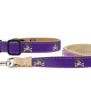 May include: A purple and tan dog collar and leash set. The purple portion features a repeating skull and crossbones logo. The collar has a black buckle. The leash has a silver-colored metal clasp.