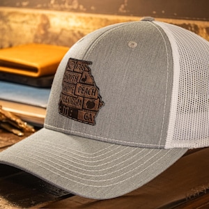 Puede incluir: Gorra de camionero gris con la parte trasera de malla blanca y visera curva. La parte delantera presenta un parche de cuero marrón con la forma del estado de Georgia, con texto como "1788", "South", "Home", "Peach" y "Georgia".