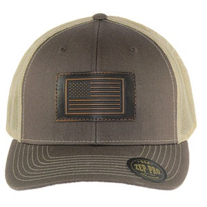 Könnte beinhalten: Eine braune und beige Baseballkappe mit einem braunen Lederpatch, auf dem eine amerikanische Flagge abgebildet ist. Die Kappe hat ein Netzrücken und ein braunes Schild mit einem schwarzen "Zep-Pro"-Etikett.